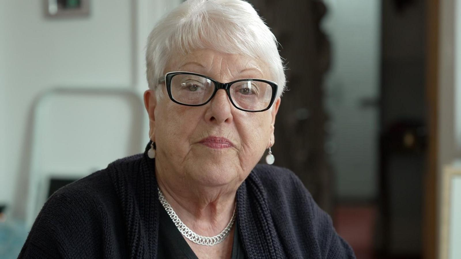 A woman with short white hair, glasses and a dark black top looks serious as she faces the camera. She is also wearing a silver-coloured necklace and earrings.