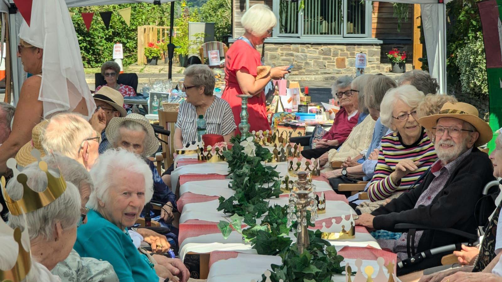 A group of older people sit at a long table which has green floral decorations on it. Some of the people are wearing gold paper crowns.