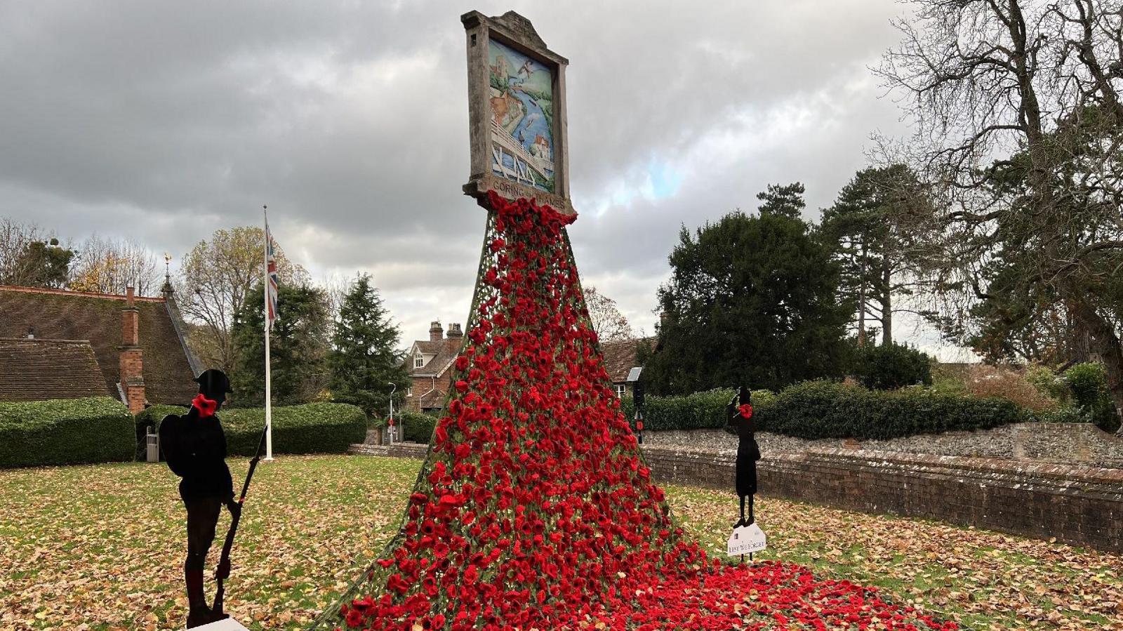 The picture shows the Goring Village sign with 1,752 hand-knitted poppies running down red camouflage netting onto the floor. 