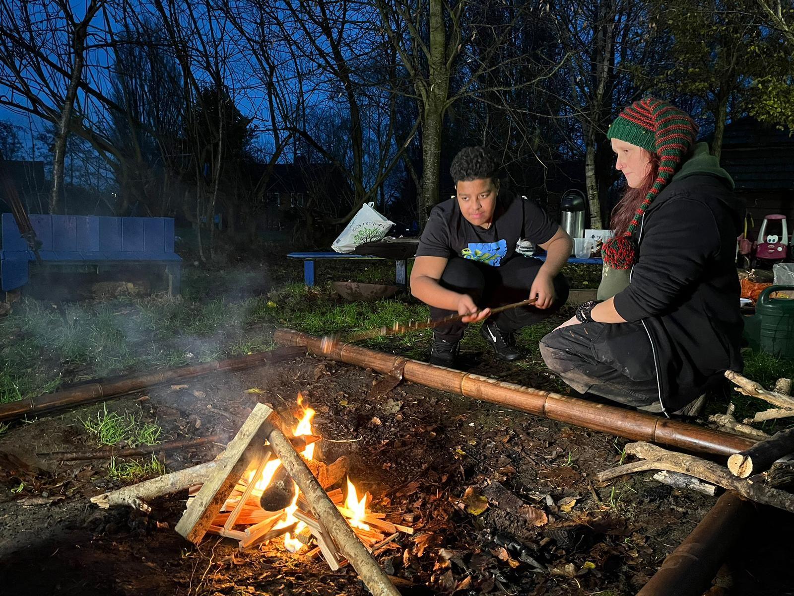 A woman wearing a woolly hat with a boy in a black t-shirt looking over a campfire 