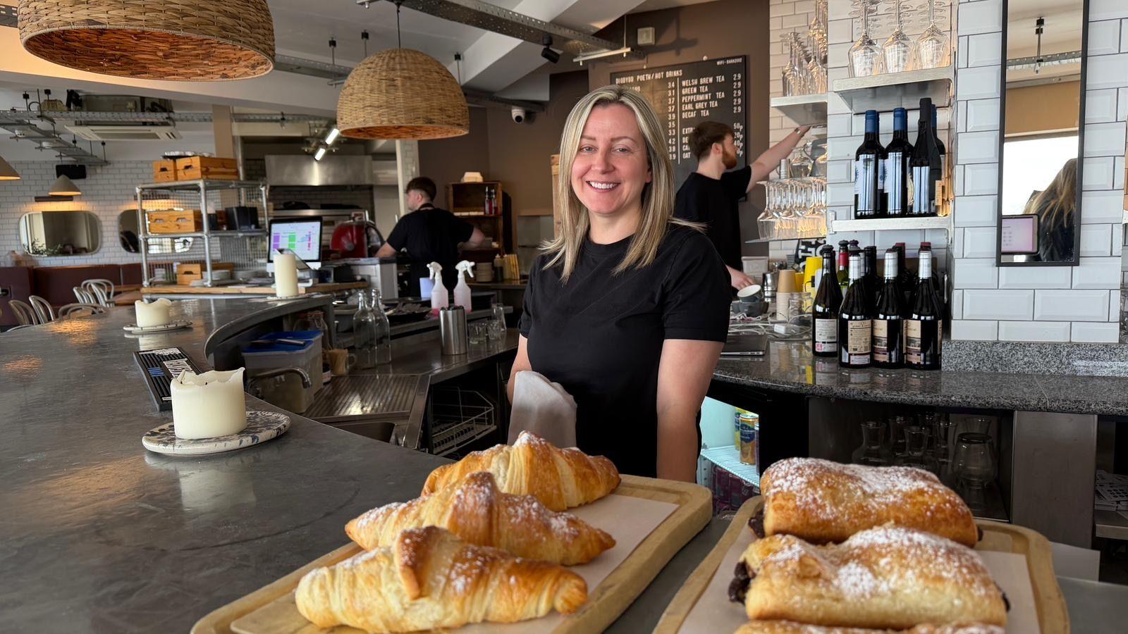 Llun o Nia Roberts wrth gownter ei bwyty - Baravin - gyda croissants ar y cownter ac aelodau staff yn parhau a'u gwaith yn y cefndir