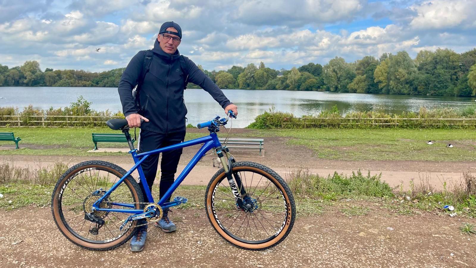 A man in stands beside a lake holding a blue bicycle. He is wearing a dark zipped hoodie, a black rucksack, glasses and a dark baseball cap worn backwards. His left hand is holding the handlebar of a bicycle. Geese and green benches can be seen in the background beside a footpath.