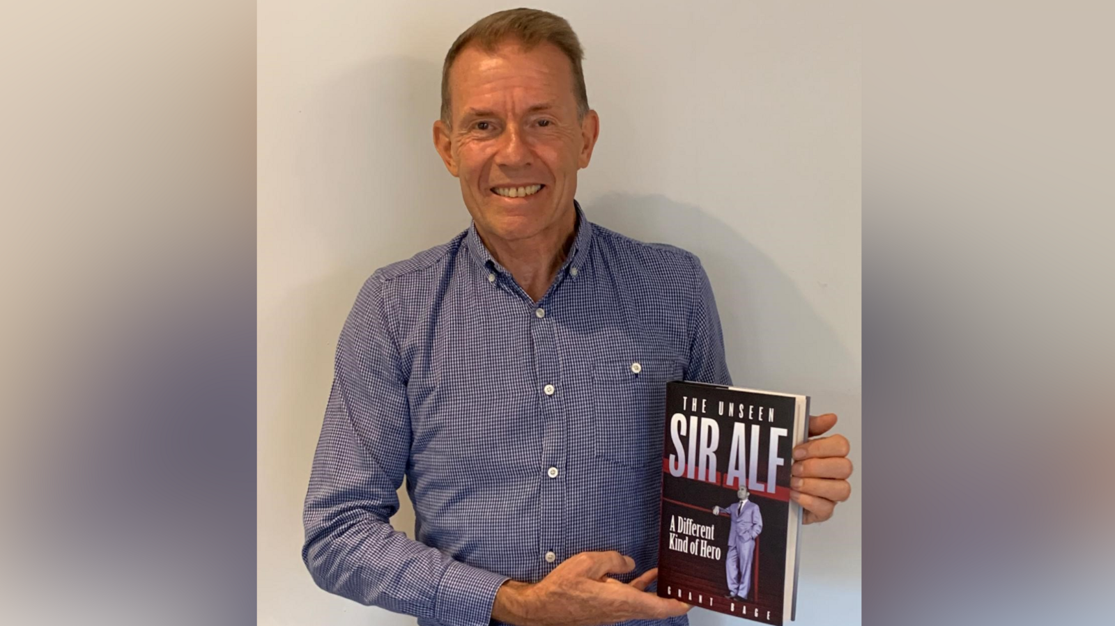 Grant Bage smiles at the camera while holding his book. He has short hair and wears a blue shirt. 