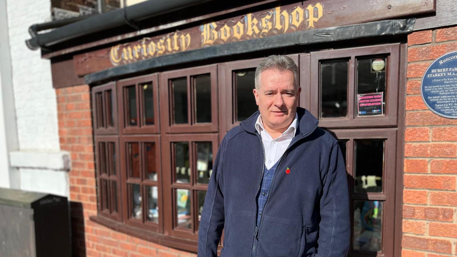 Chris Howard, in a blue zip-up fleece, stands outside his bookshop with a sign reading 'Curiosity Bookshop'