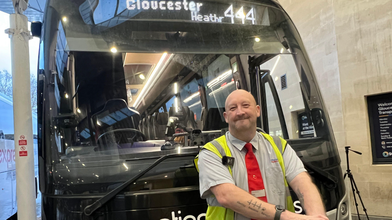 A coach driver stands in front of his 444 Gloucester to Heathrow National Express route. He is bald and is wearing a grey short sleeved shirt, a red tie and a yellow hi-vis tabard. He is smiling and showing off a 444 tattoo he has on his forearm, which has writing tattooed beneath it.