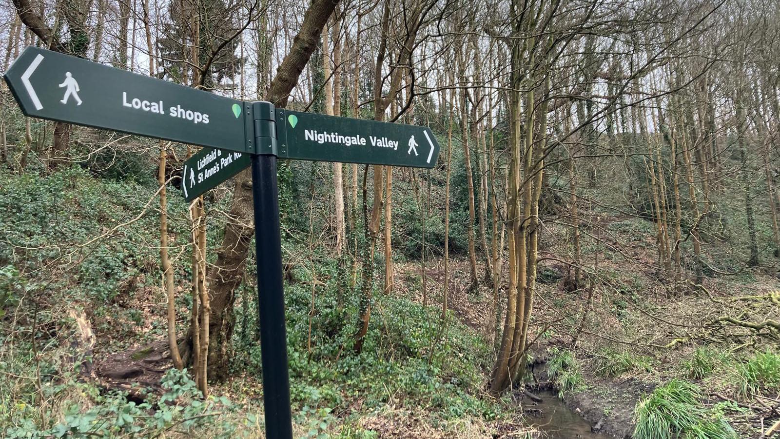 A signpost in the woods. One pointing towards Nightingale Valley and another pointing towards local shops.