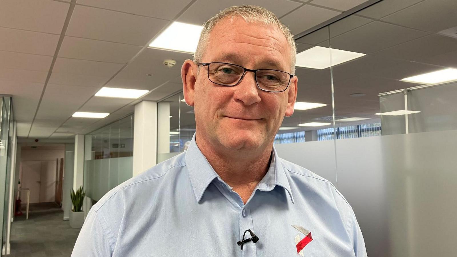 A man stands in a corridor in an office building. The man has grey hair, and he is wearing glasses and a light blue shirt with a red and white company logo on it.