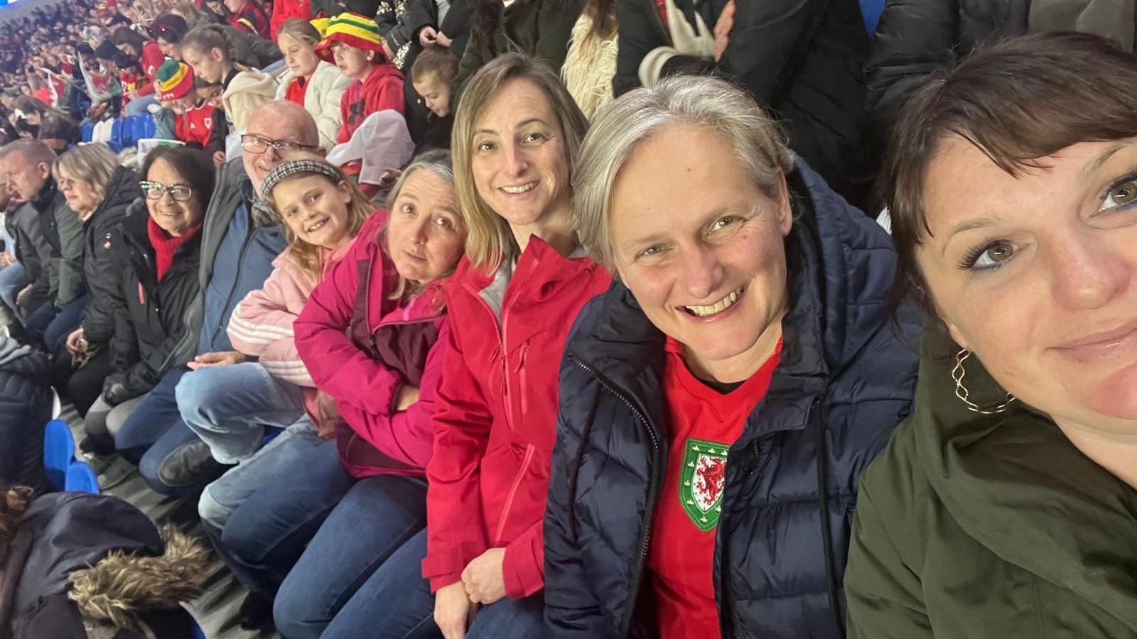 A selfie of fans at the football. Ruth is on the far right and is taking the photo, she is wearing a green jumper and has gold hoop earrings and is smiling. A woman is sat next to her and is wearing a red Wales shirt, she has grey hair and is also smiling. Next to her is a woman with blonde hair and a pink jacket. She is next to another woman with a pink jacket. Next to her is a girl with a pink Adidas coat, who has her arms crossed and is smiling at the camera. Next to her is an older couple who are both smiling at the camera.