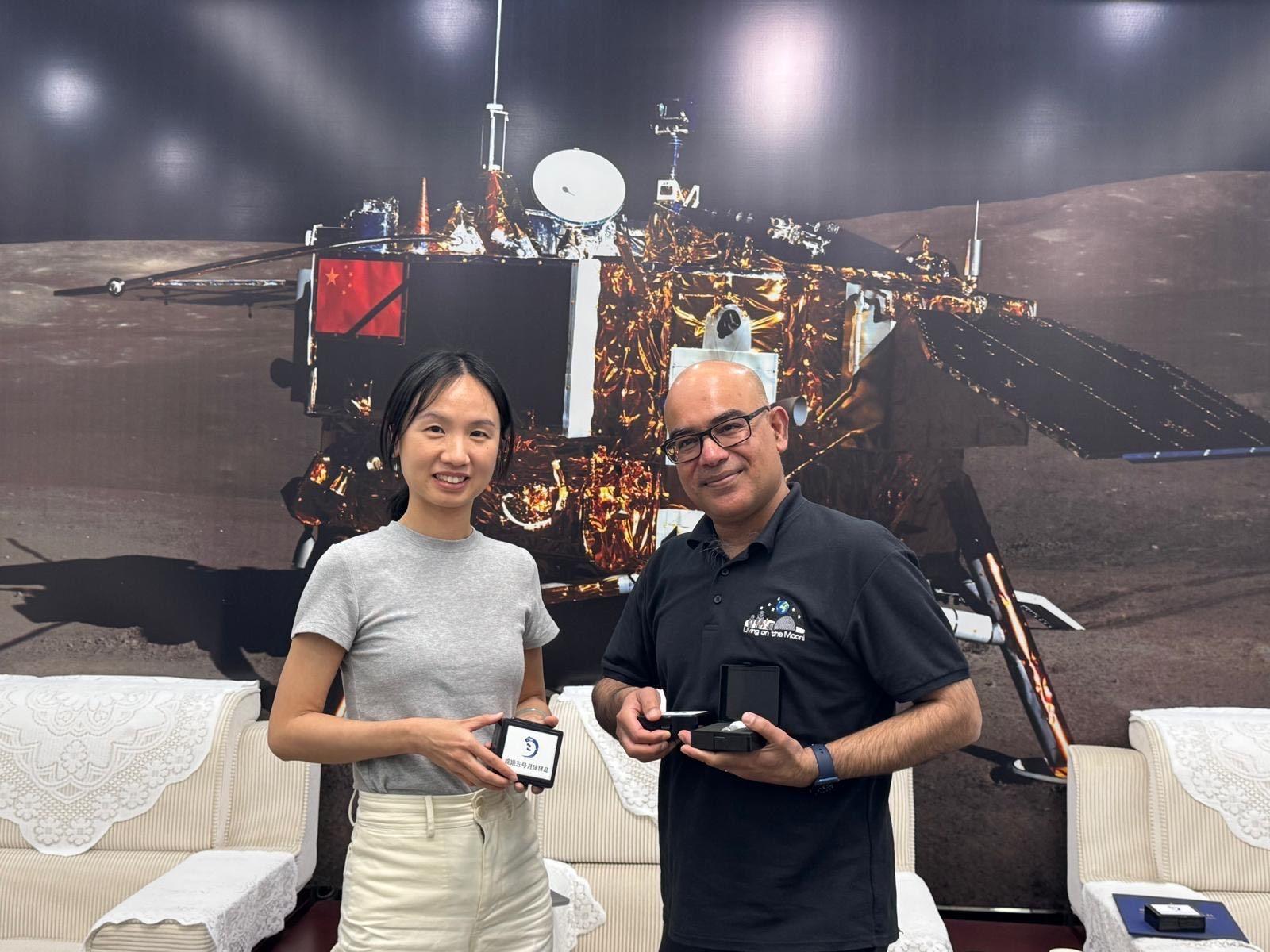Mahesh Anand and a woman working with the Chinese space agency standing in front of a large picture of a spacecraft with black arms and gold material on the outside. They are standing in front of white sofas and holding small boxes. The boxes have white labels with the Chinese space agency logo.
