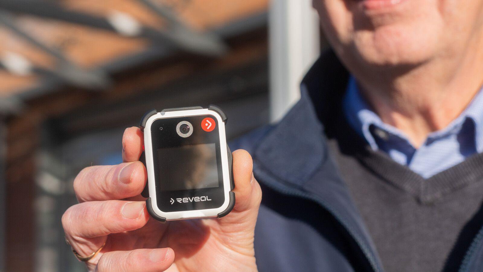 A person holds a small square-shaped bodycam device, featuring a screen and camera lens on the front. The individual is dressed in a dark jacket over a light blue collared shirt.