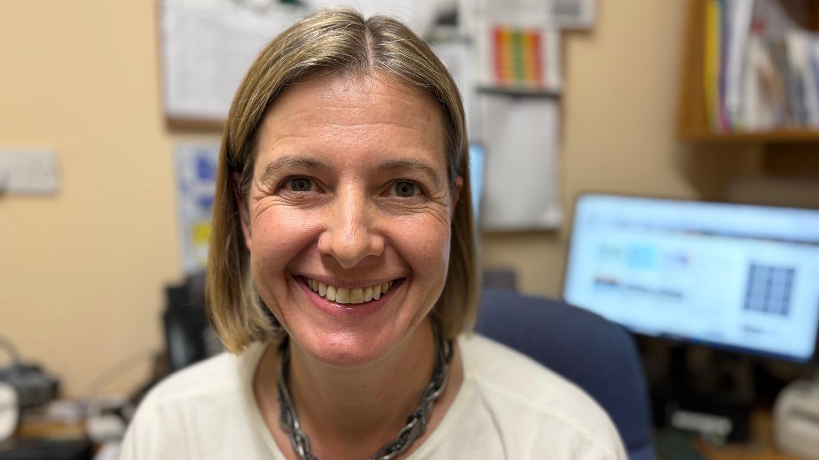 Dr Davies is sat with her desk in the background. She is wearing a white jumper and metallic necklace. She has chin-length fair hair and is smiling at the camera.