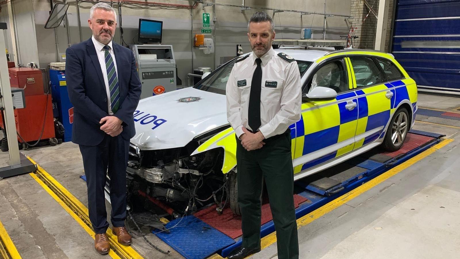 Liam Kelly and Bobby Singleton stand in front of a wrecked police car. The car has blue and yellow police colours, with a white bonnet. Their is a chunk taken out of the front left of the car, inbetween the grill and the left wheel. Inside the car airbags have been deployed. Liam Kelly stands on the left in a navy suit, white shirt, brown shoes and a green and navy striped tie. Singleton wears his police unifrom, which consists of green trousers, a white shirt, black shoes  and a black tie. 