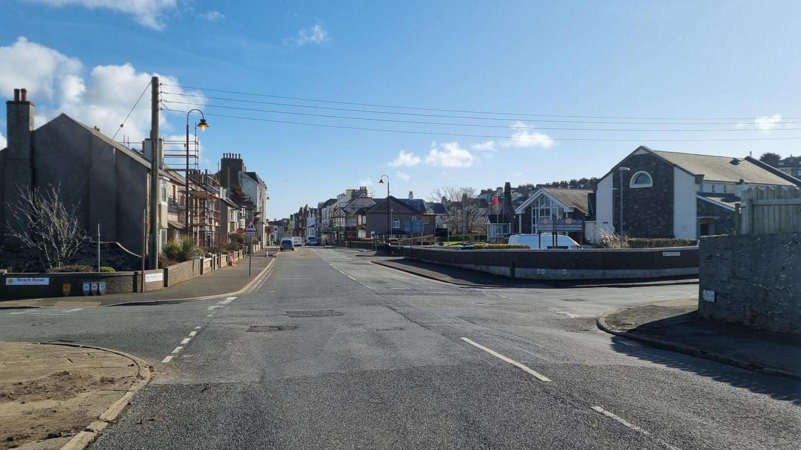 An empty junction at a roadside. A large school building can been seen next to it. Houses line the other side of the road. The sky is powder blue with a few wispy clouds.