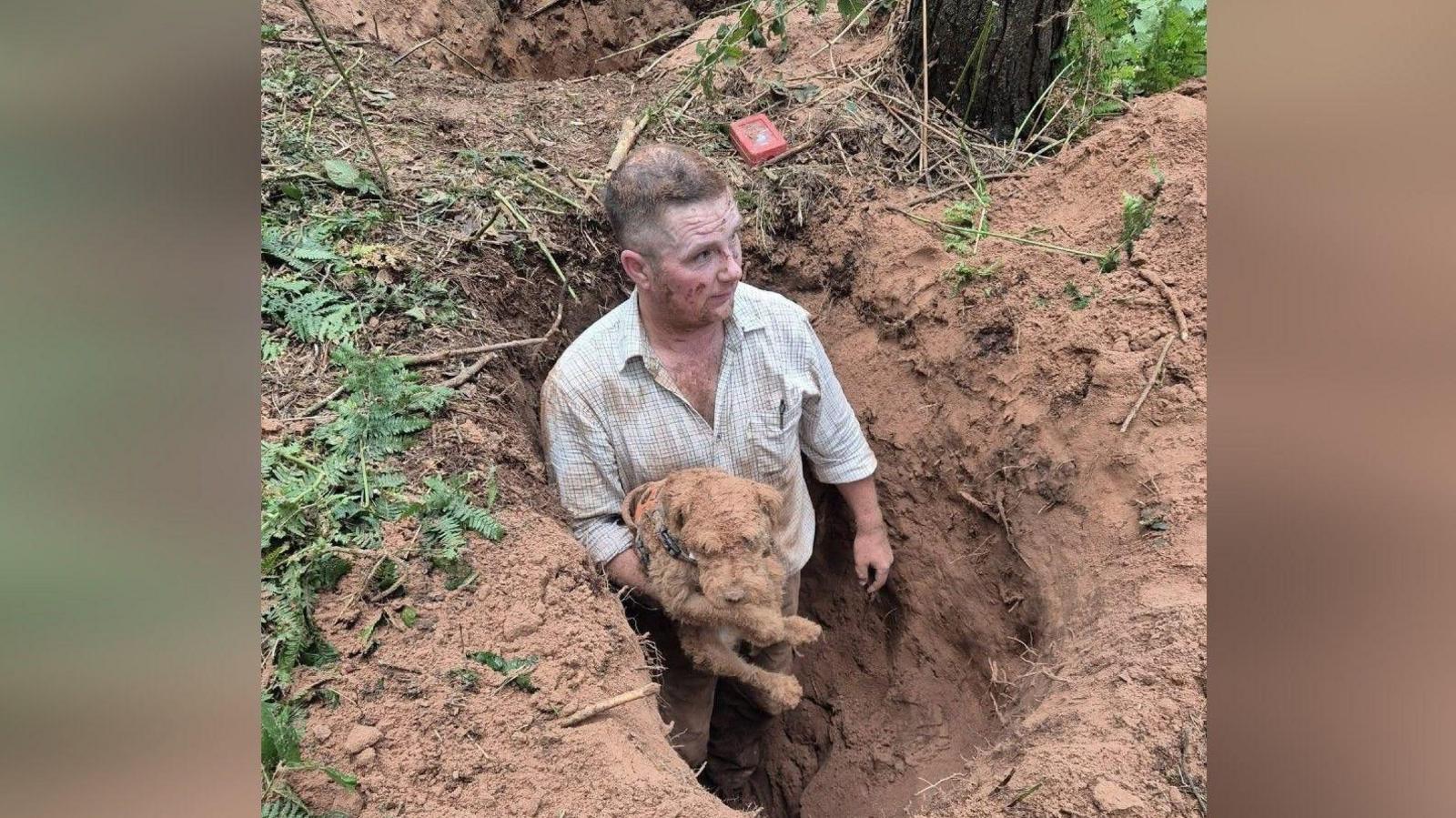 Gordon Bottomley coming out of a hole in the ground holding Sherlock the Lakeland terrier. Mr Bottomley is wearing a light chequered shirt and brown trousers and he covered in dust, including on his face and clothing. He is standing at the entrance of a narrow underground tunnel, which is as tall as the length of his body, from his shoulders to his toes. Sherlock is in his arms and he is the same colour as the sandy soil surrounding them.
