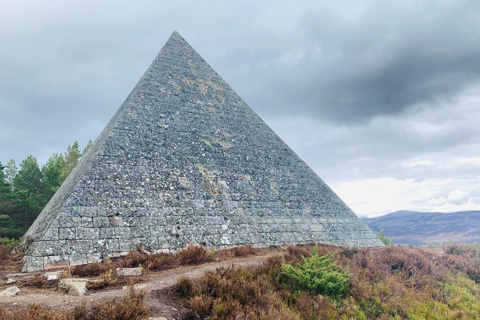 Pyramid-shape cairn at the edge of woodland, with hillside and a cloudy grey sky in the background.
