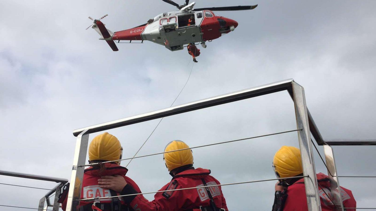 Three lifeboat crew members, in red uniforms and yellow helmets, watch from a platform as a helicopter winches a casualty from Spitbank Fort.
