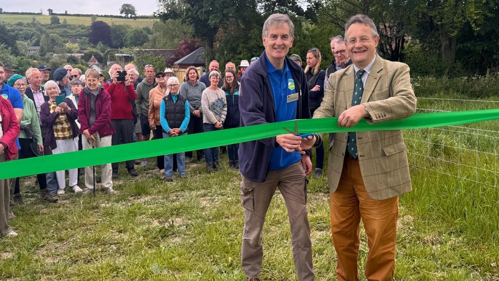 Two men, one in a brown tweed suit, another in a blue polo top and navy hoodie, holding a pair of scissors and cutting a green ribbon. There is a crowd of people behind them smiling and taking photgraphs. They are all standing outside in a field.