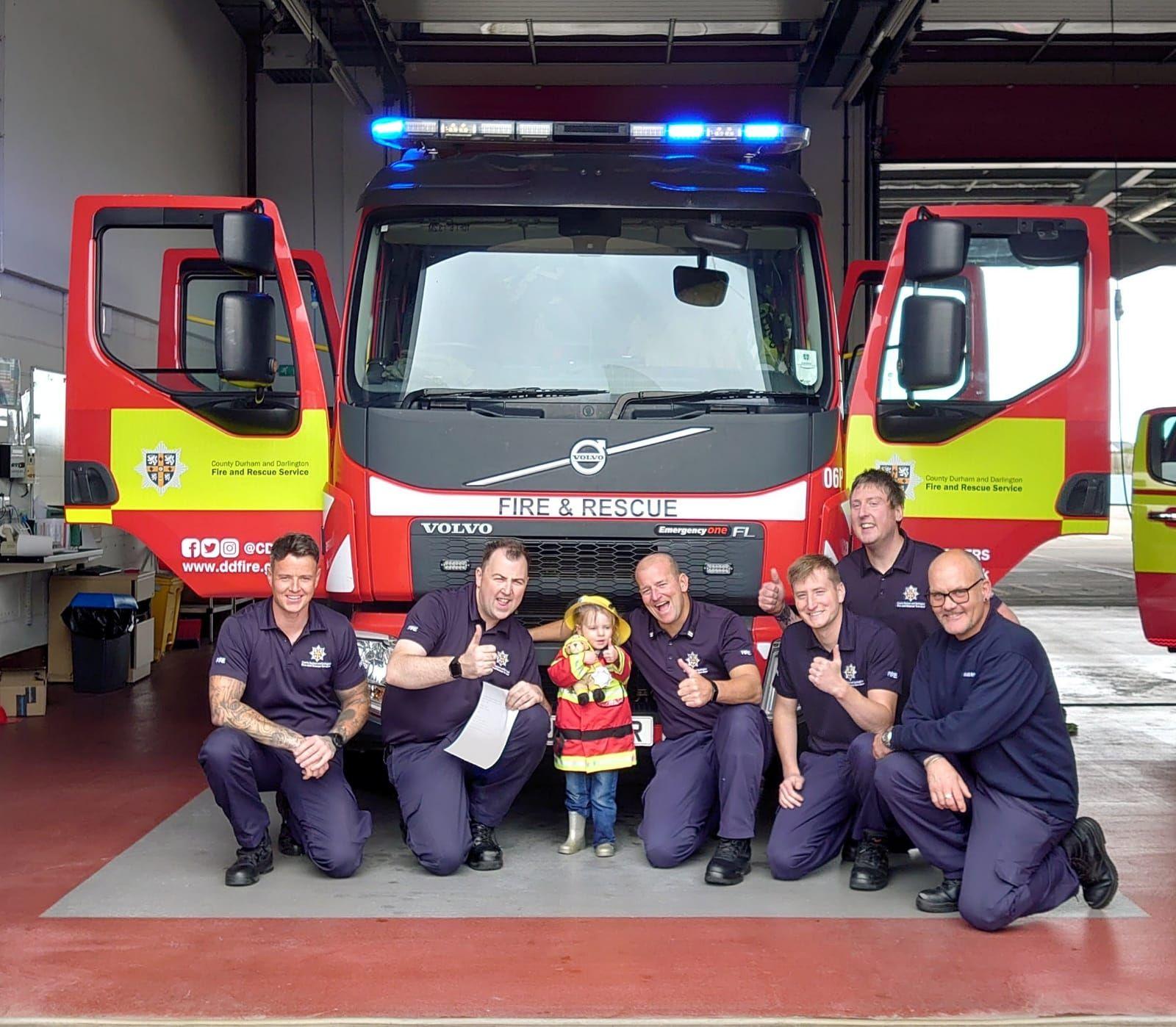 Beatrix and a group of firefighters pose in front of a fire truck. The blue lights of the truck are on. They are all smiling at the camera, some have their thumbs up.