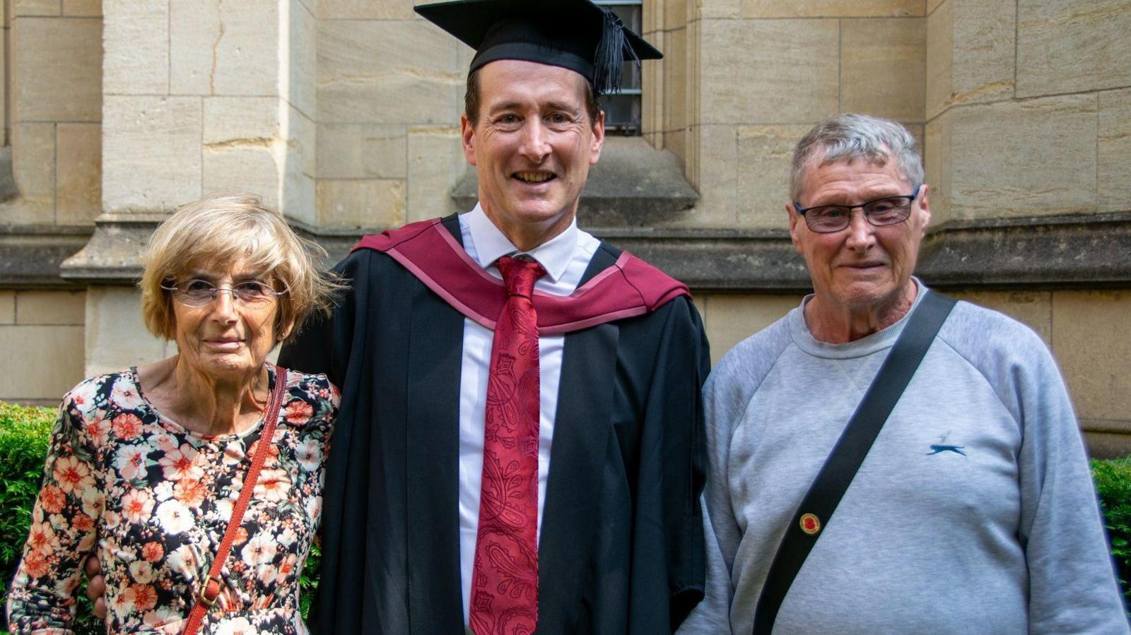 Mathew standing in the middle of his parents at his graduation day