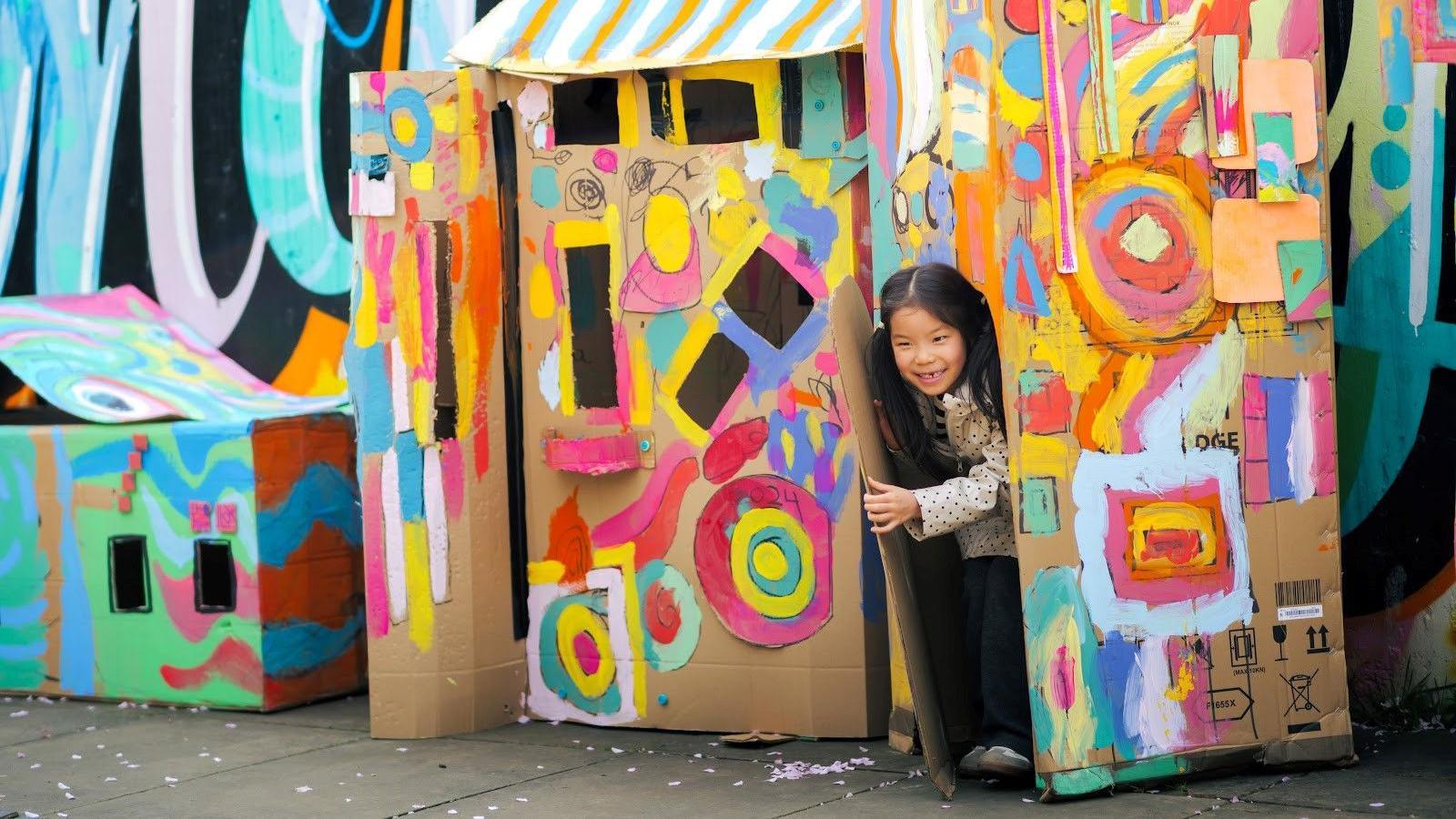 A little girl with long black pony tails opens the door of a cardboard house painted with colourful shapes.