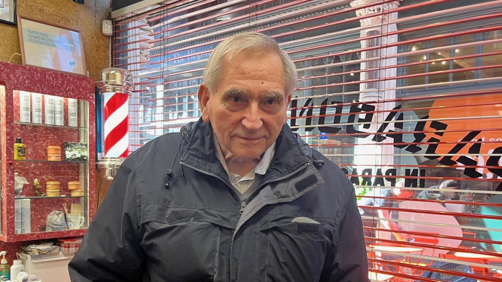 Gordon Readhead, 81, with grey hair and thick eyebrows, is wearing  a grey and standing in the shop, with the window behind him. The window has open, red blinds.