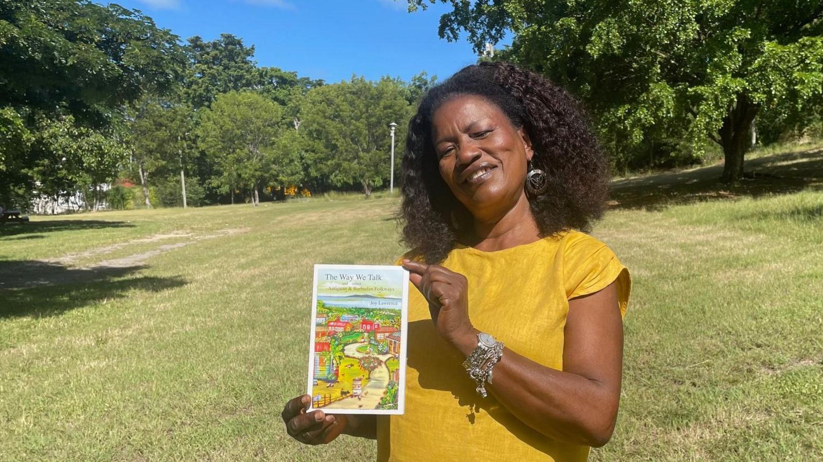 Joy Lawrence, surrounded by grassland and trees, holds up her book The Way We Talk