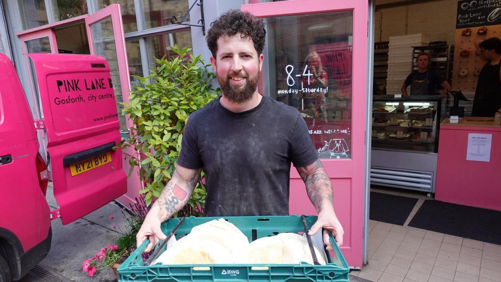Abe Chirgwin is standing outside Pink Lane Bakery holding a green box full of stotties. The bakery doors and counters are pink. Mr Chirgwin has dark curly hair and a beard and is wearing a grey t-shirt.