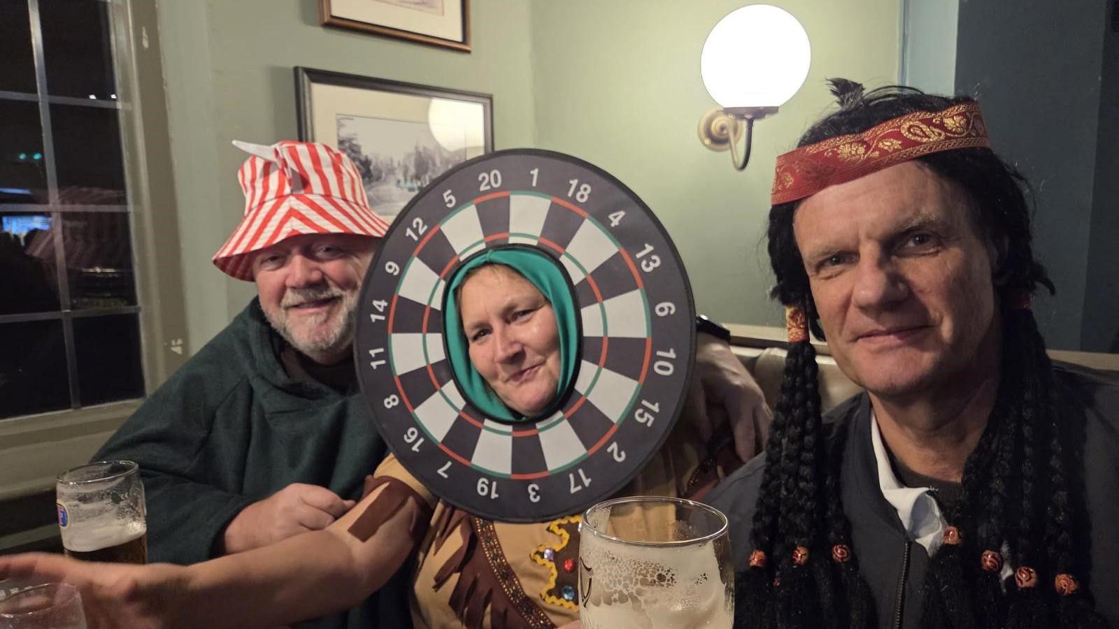 Three people are dressed up, including the man on the left who is wearing a red and white hat. Next to him is a dartboard, which has someone's head in the middle of it.