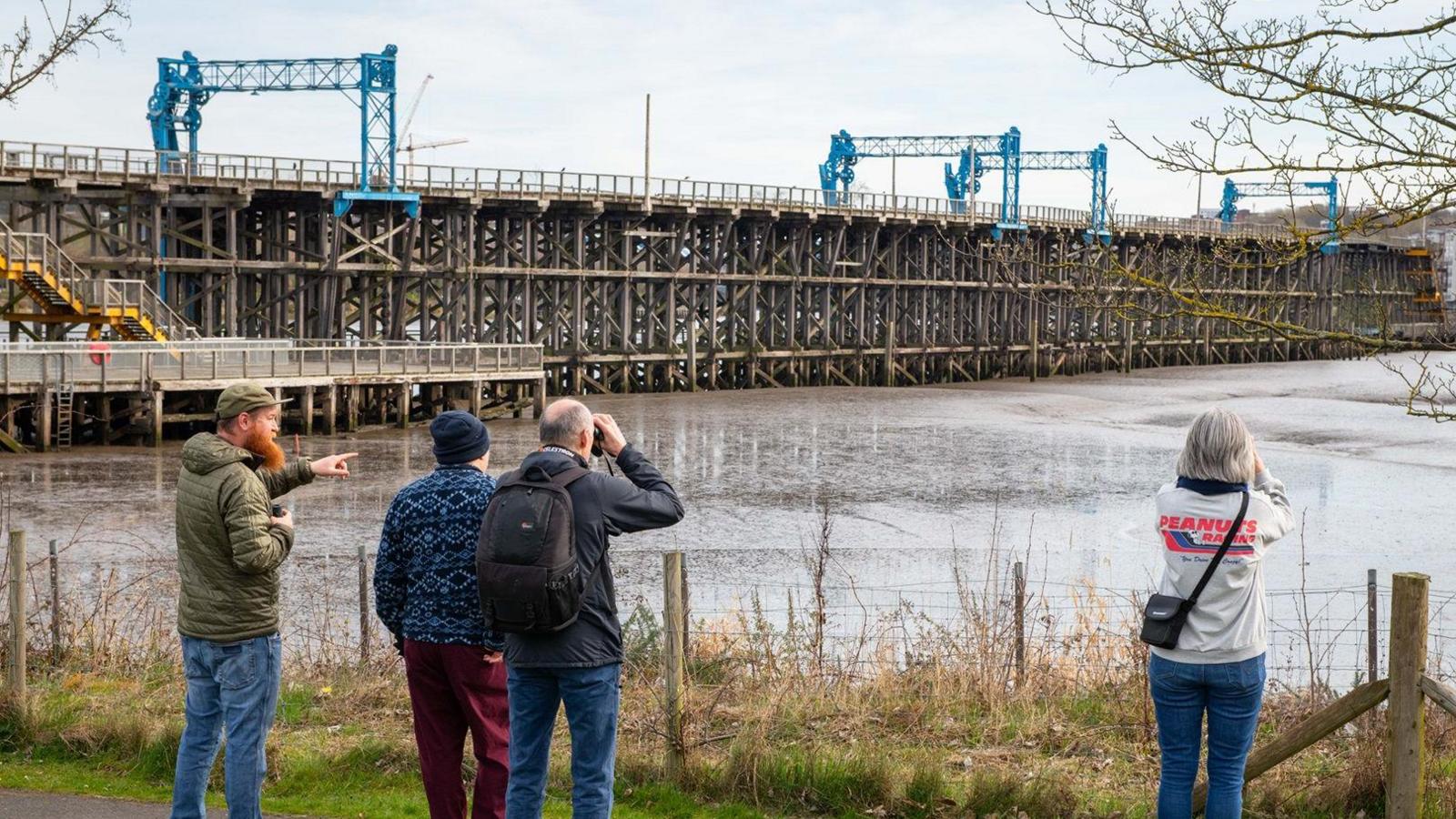 Four people, two using binoculars, are pictured from behind, looking out from a path towards Dunston Staiths and the tidal basin. Dunston Staiths is a wooden structure similar in appearance to a pier. 