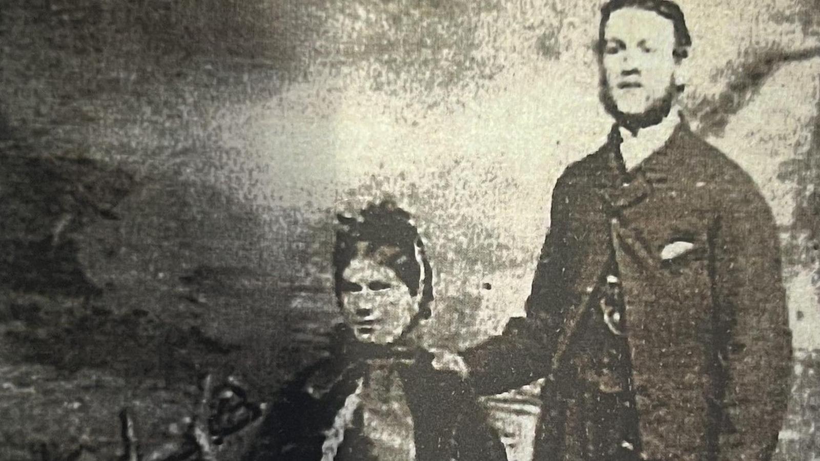 A faded black and white photo of a smartly dressed man and woman. She is sitting down and he stands with one hand on her shoulder. They are dressed in Victorian era clothing.
