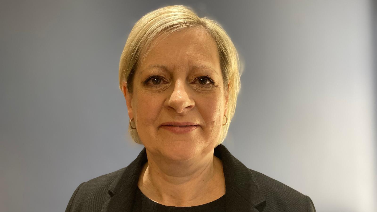 Professor Karen Dunderdale is pictured portrait style against a grey wall, she has short blonde hair and is wearing a black top and jacket