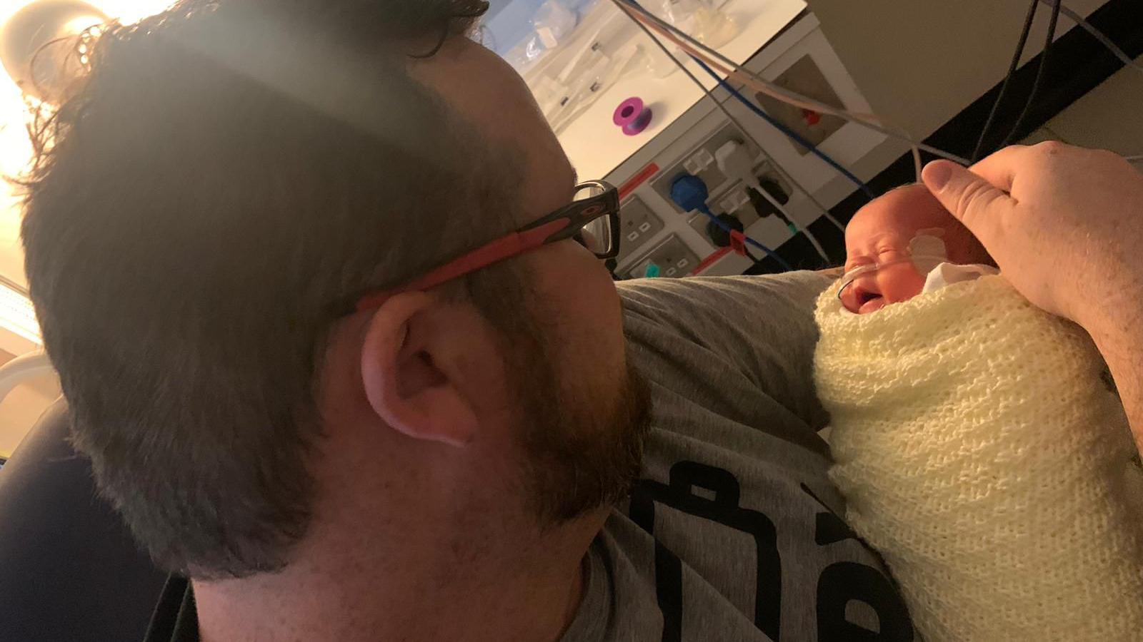 Jon holds baby Ralphie in his arms. The baby is wrapped in a blanket and has an oxygen tube taped to his nose. There is medical equipment behind them.