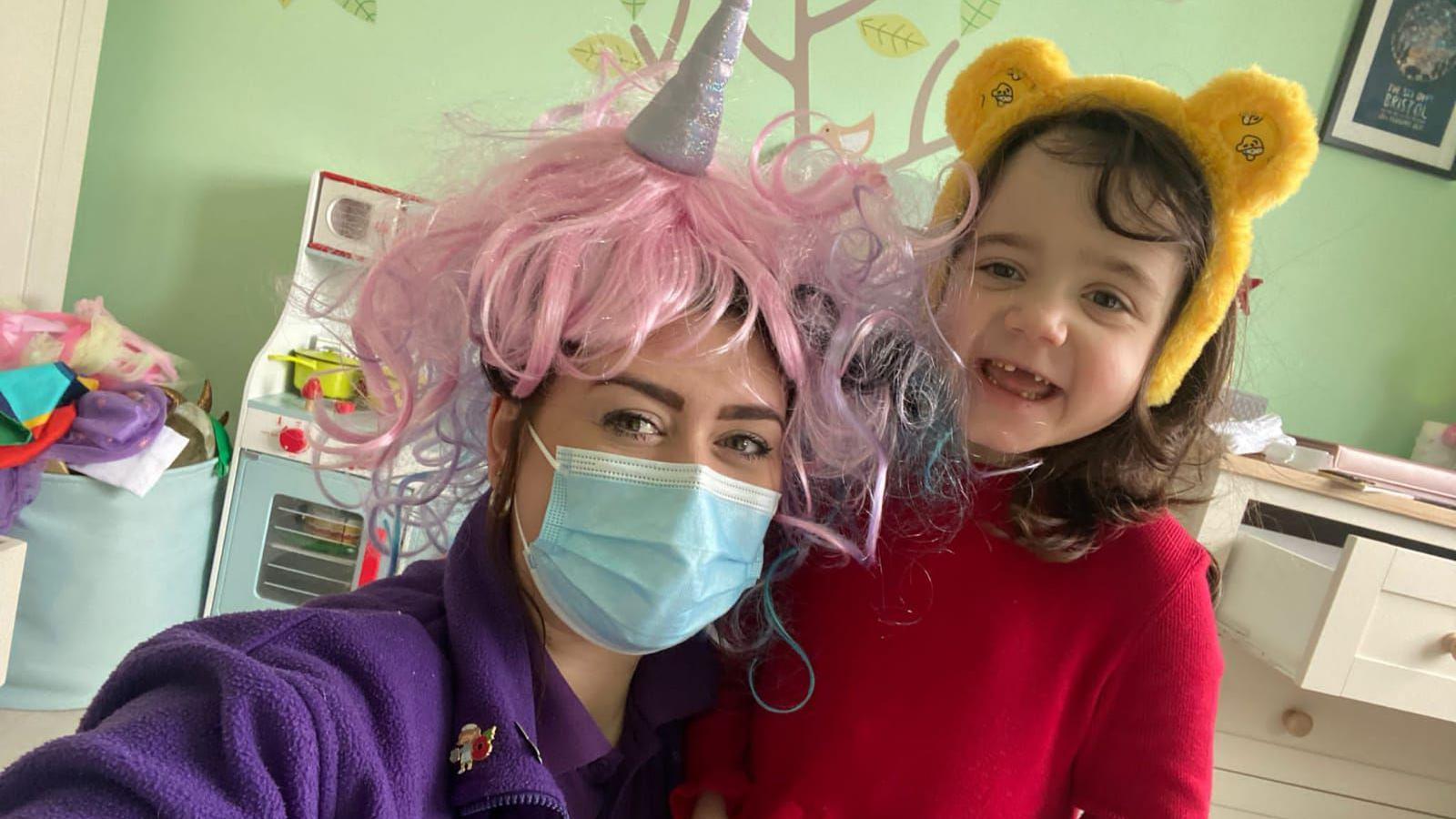 Izzy, wearing a Pudsey teddy bear set of ears, and her nurse Charlotte in a purple jumper and pink wig in a hospital room