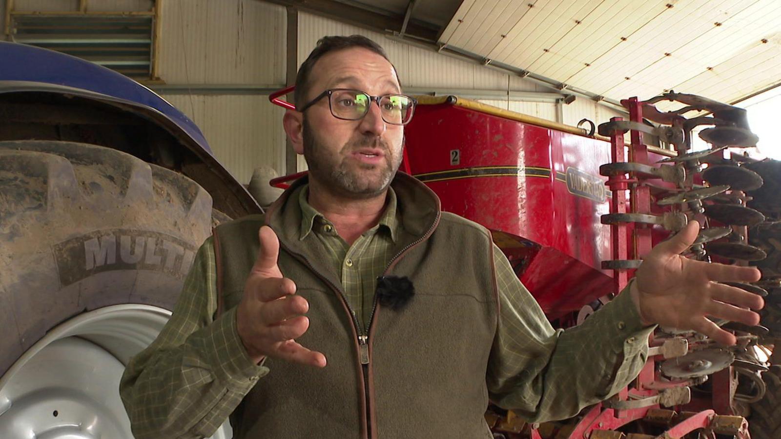 Andy Shaw, with brown hair and glasses and wearing a green gilet over a green cheque shirt, speaks to the camera in front of a tractor and farm equipment. 