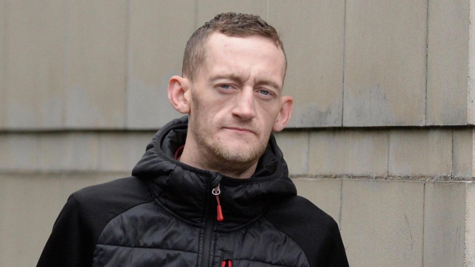 Craig Rowland - a man looks at the camera as he stands outside a white-stone building. He has short brown hair and is wearing a black puffed jacket with a red zip.