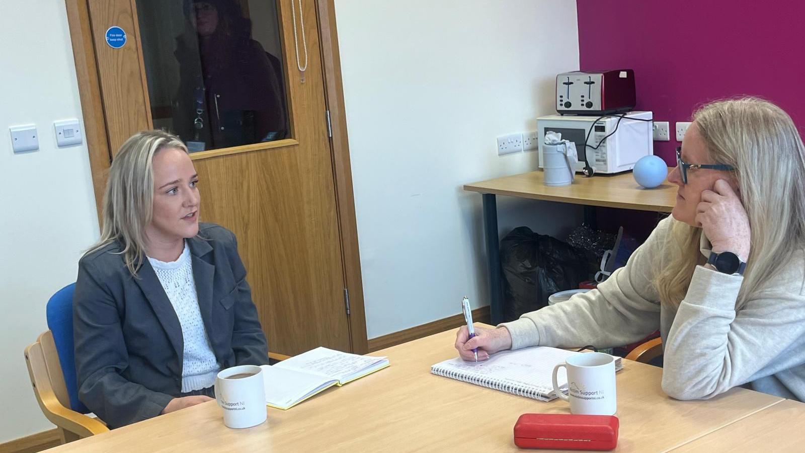 chloe wilson and Geraldine hanna sit at either side of a desk in an office. chloe is on the left wearing a grey jacket and white top. Geraldine is holding a pen and writing on a note pad. both have cups of coffee in front of them. a 