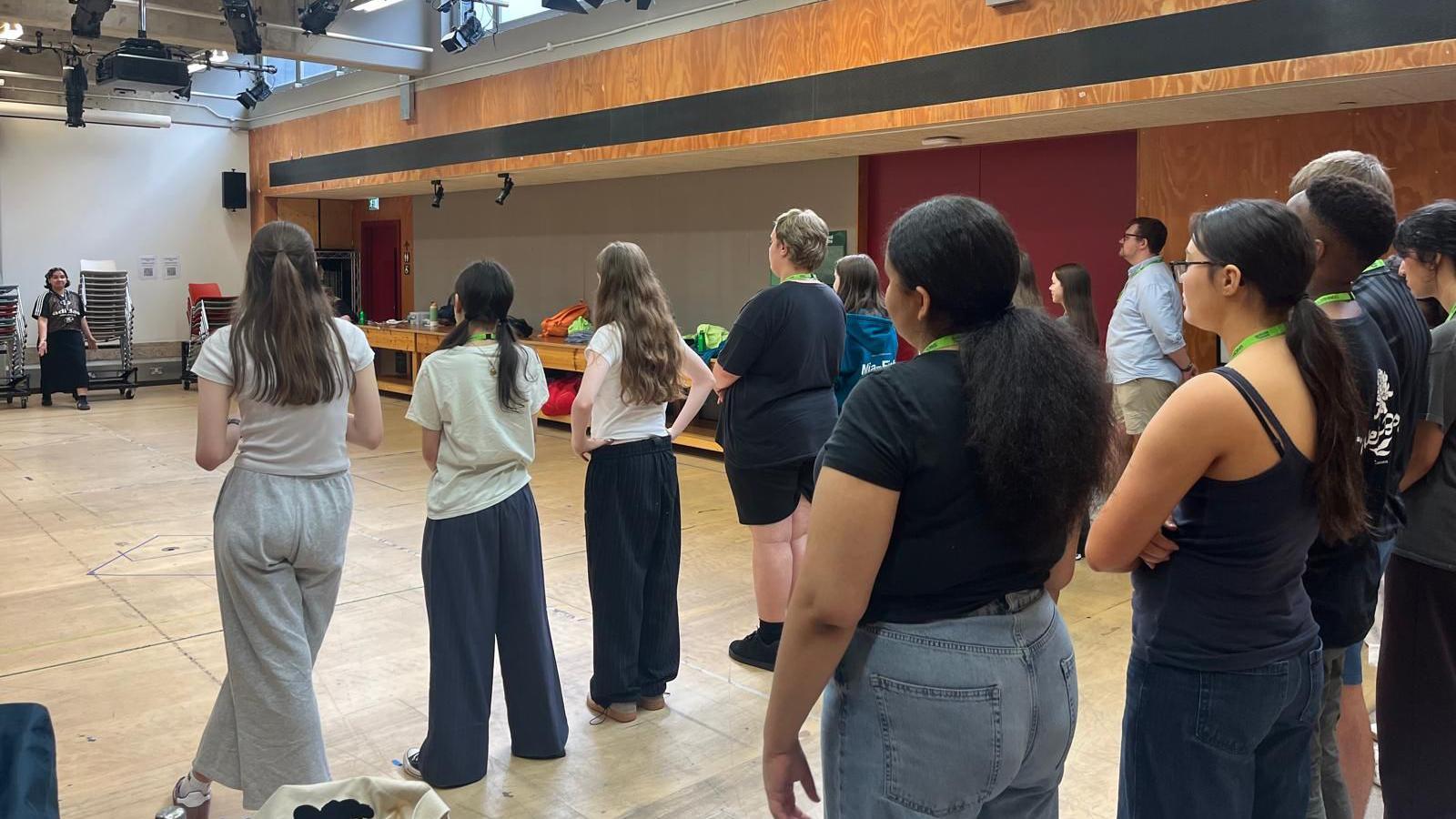 School students in a rehearsal room. They are standing in rows on a wooden floor, watching a teacher at the other end of the room.