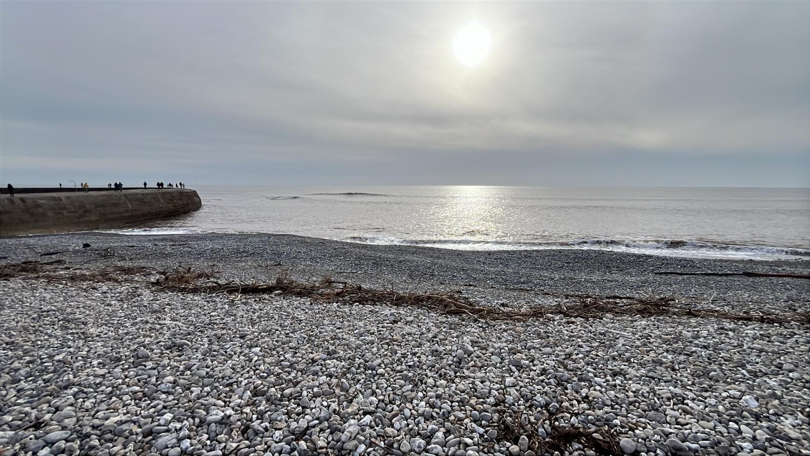 Looking out to see across a silver/grey pebble peach at Lyme Regis, with the harbour jutting into the ocean on the left. A line of seaweed shows where the tide turned, and a small wave crashes. The sun is lower in the sky and casting a white/yellow shine across the sea between itself and the shore.