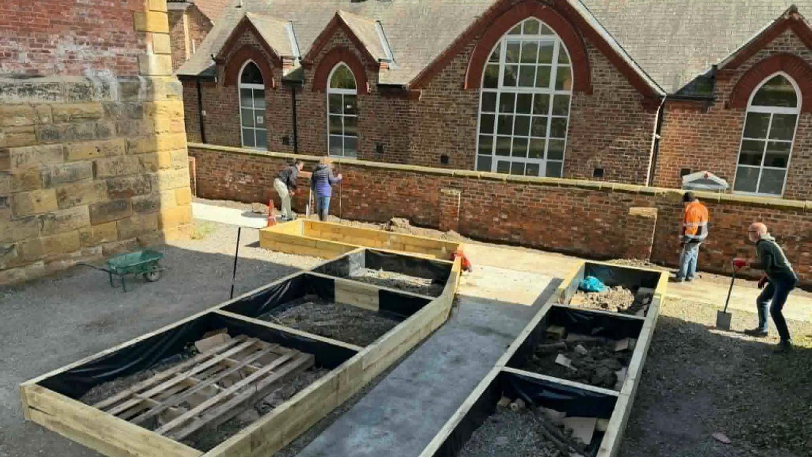 Four men and women using gardening tools to create a raised wooden flower beds area. There is an old stone building in the background, a brick building with four large windows and a brick wall.