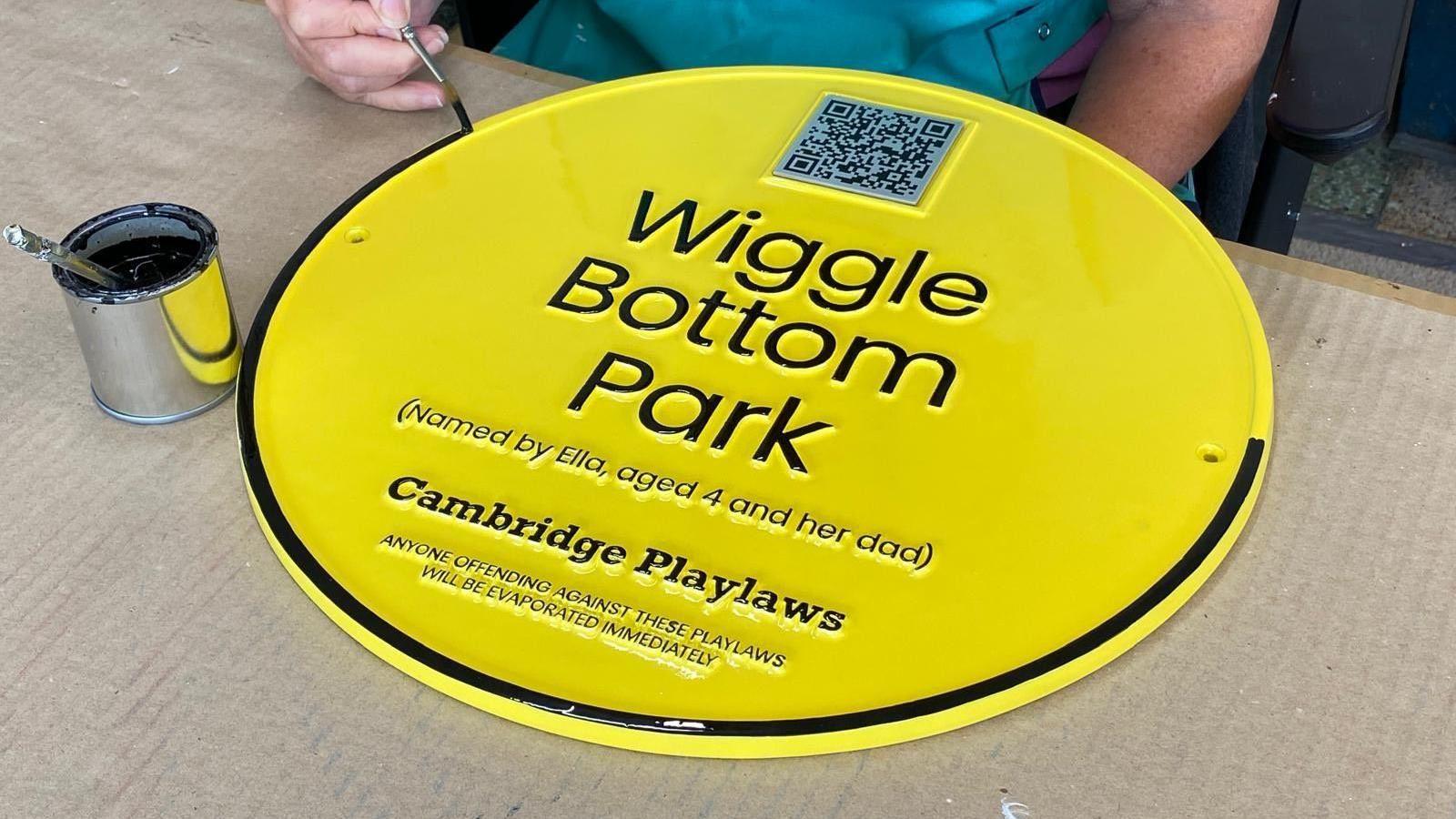 A bright yellow plaque being painted with a black outline. The plaque says: "Wiggle Bottom Park (named by Ella, aged 4 and her dad), Cambridge Playlaws, anyone offending against these playlaws will be evaporated immediately.