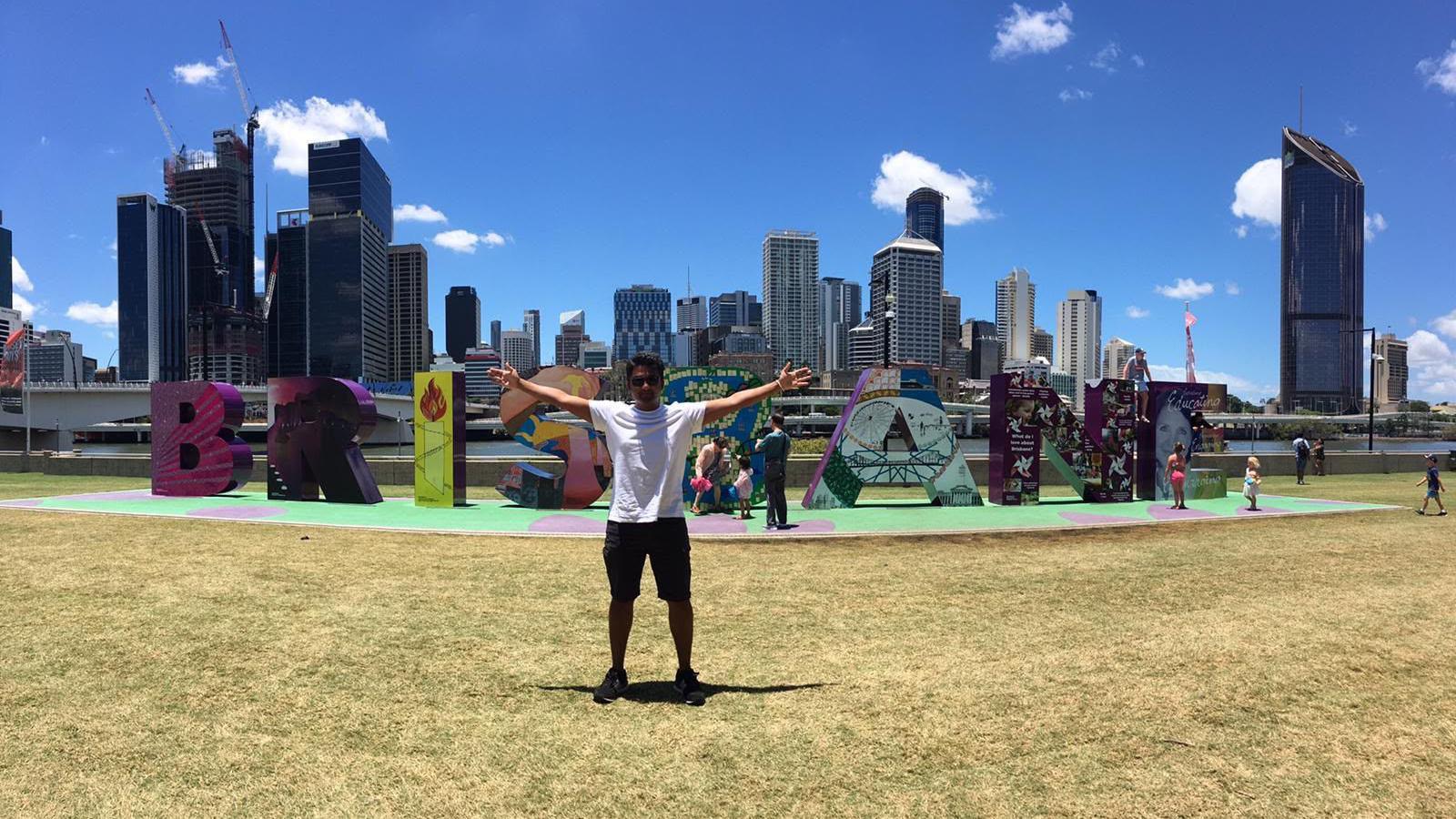Korambayil stands with his arms stretched out in front of giant letters spelling Brisbane. He is wearing a white t-shirt and black shorts.