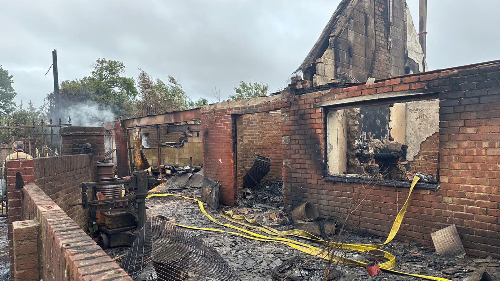 The burnt out shell of a red brick built property. It is extensively fire damaged and almost completely destroyed with only some external walls still standing, parts of the house are still smouldering and blackened debris is all over the floor.