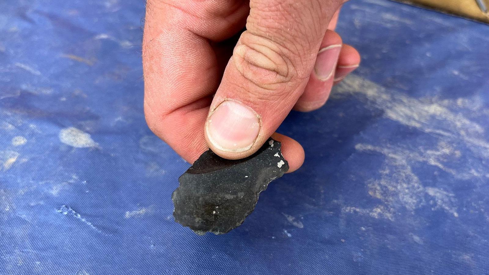 A hand holding a small, sharpened piece of black flint over a blue background