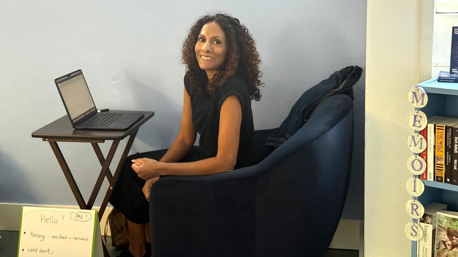 Kathryn Croft, a woman with long dark curly hair, wears a black sleeveless top and sits at a laptop on a desk in the Paper Moon bookshop in Guildford, smiling into the camera.