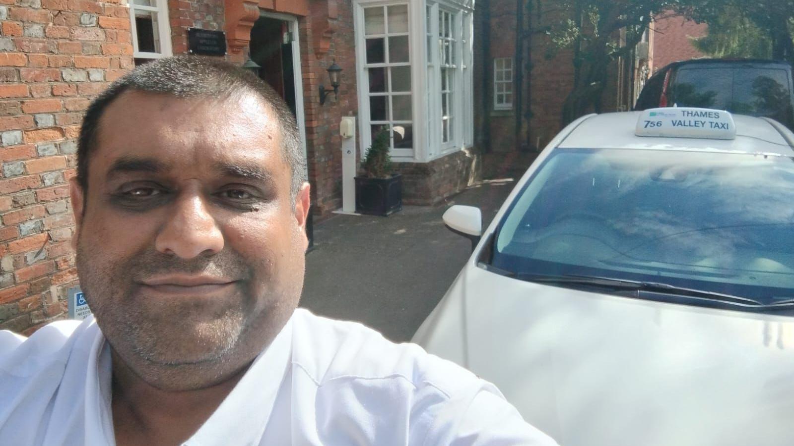 Yaseen Yusuf smiles for a selfie outside his home. He has a shaved head and light beard. He is wearing a smart white shirt and his white cab can be seen in the background.