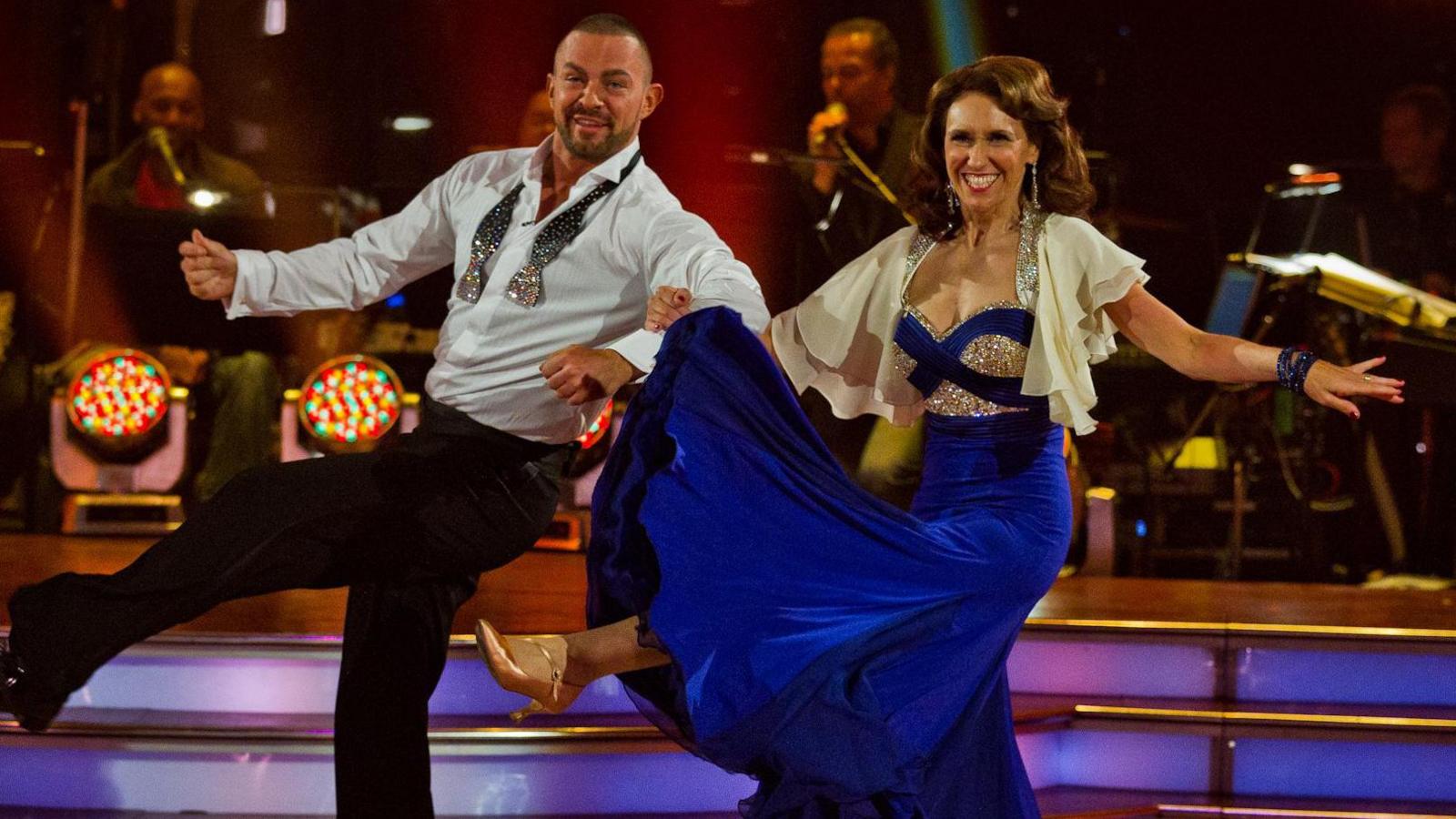 Man and woman in glamourous outfits dancing and smiling. Woman wears silver, royal blue satin dress with black velvet brocade round the bust and a flouncy satin cream bolero jacket. Man wears pale blue shit with silver 