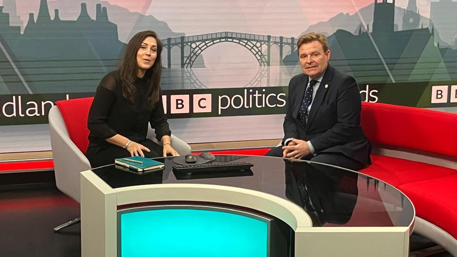 A woman with long dark hair, wearing all black, sits on a grey swivel chair with a red cushion. Opposite her is a man with light brown reddish hair, wearing a black suit, light blue shirt, dark spotted tie and with a badge on his lapel. He is sat on a long red and grey sofa. There is a curved desk in front of them. Behind is a mural with a bridge and some buildings and below it it says BBC Politics.