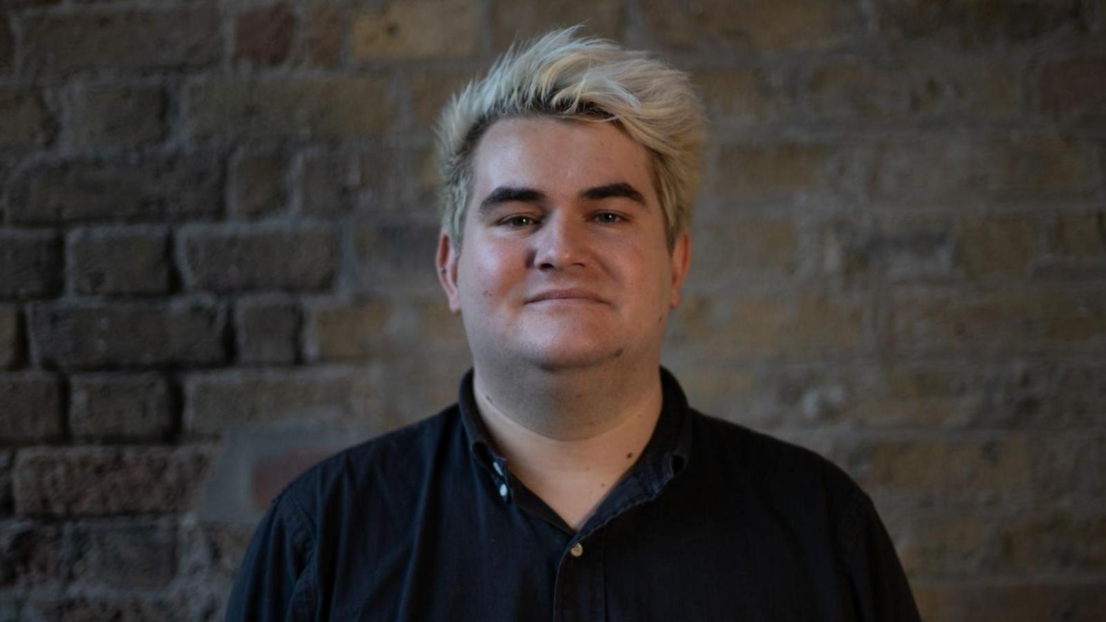 Jake Hurfurt looks directly at the camera. He has died blond hair with black roots showing, is clean shaven and wears an open collar black shirt. Behind him is an exposed brick wall.