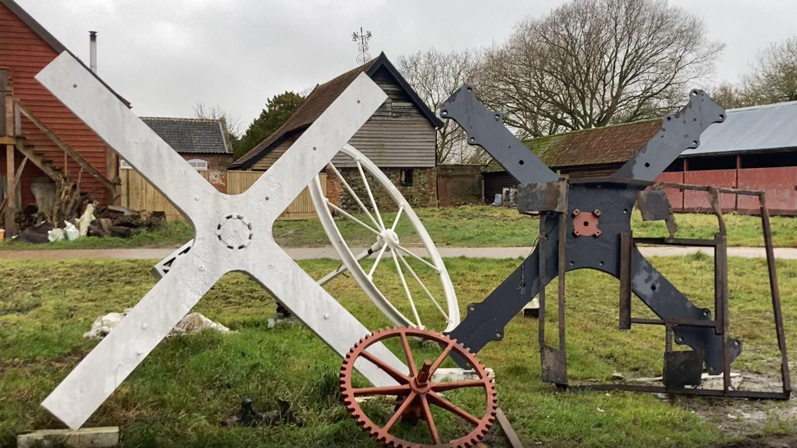 A large white part of a windmill and a smaller black one next to it sit outside several buildings on grass. They are in the shape of a cross. Other wheel parts sit in front and behind them.