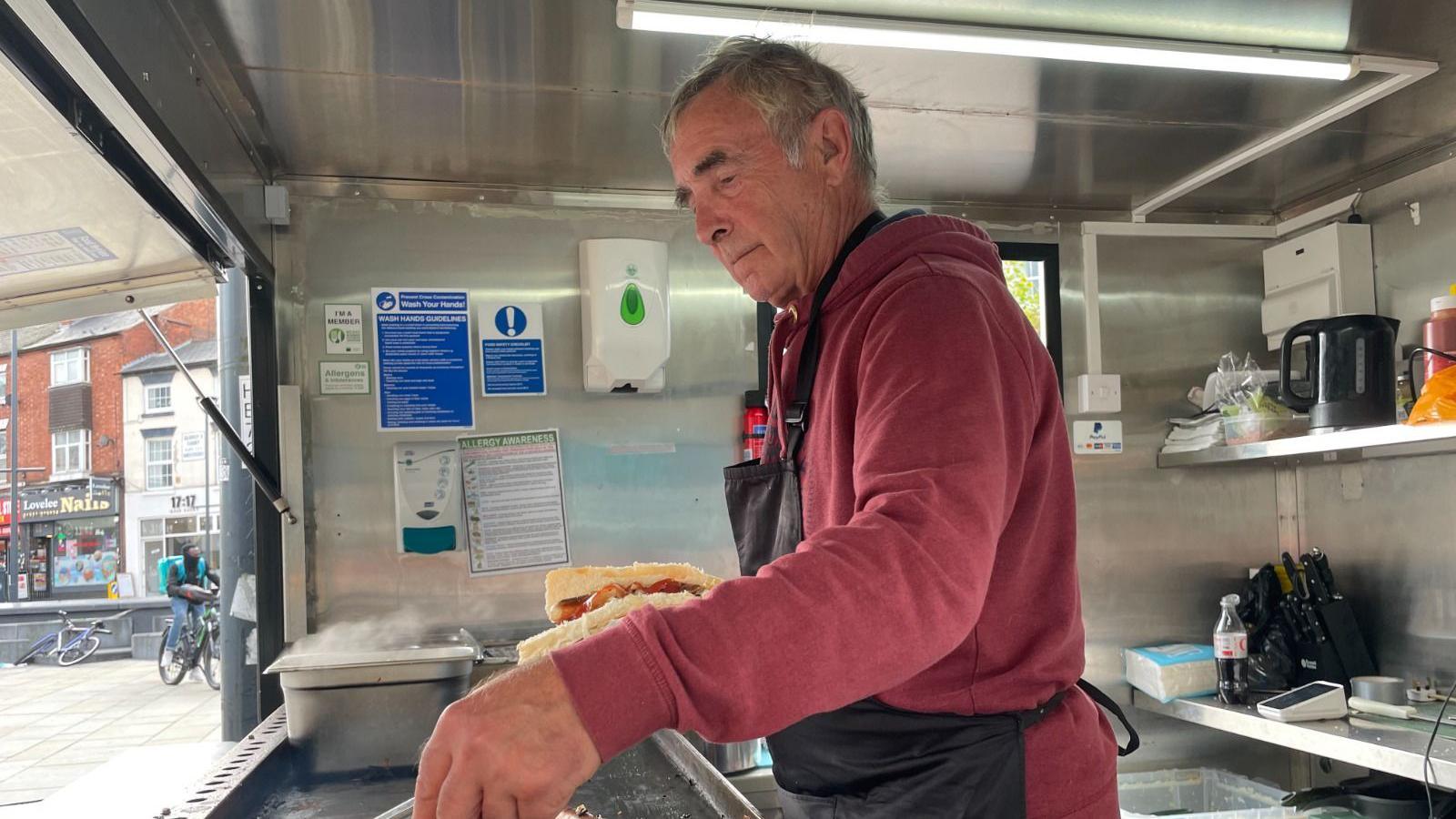 Charlie Pearson wears a red hoody and apron and is cooking hot dogs on a hot plate.